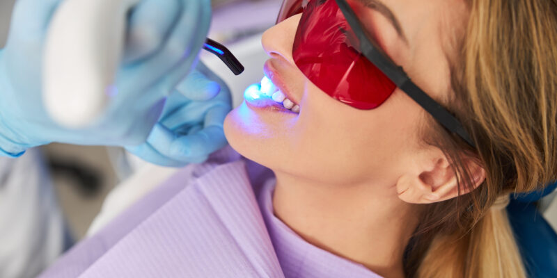 Woman covering her eyes with red plastic glasses and smiling while undergoing LED curing lights dental treatment